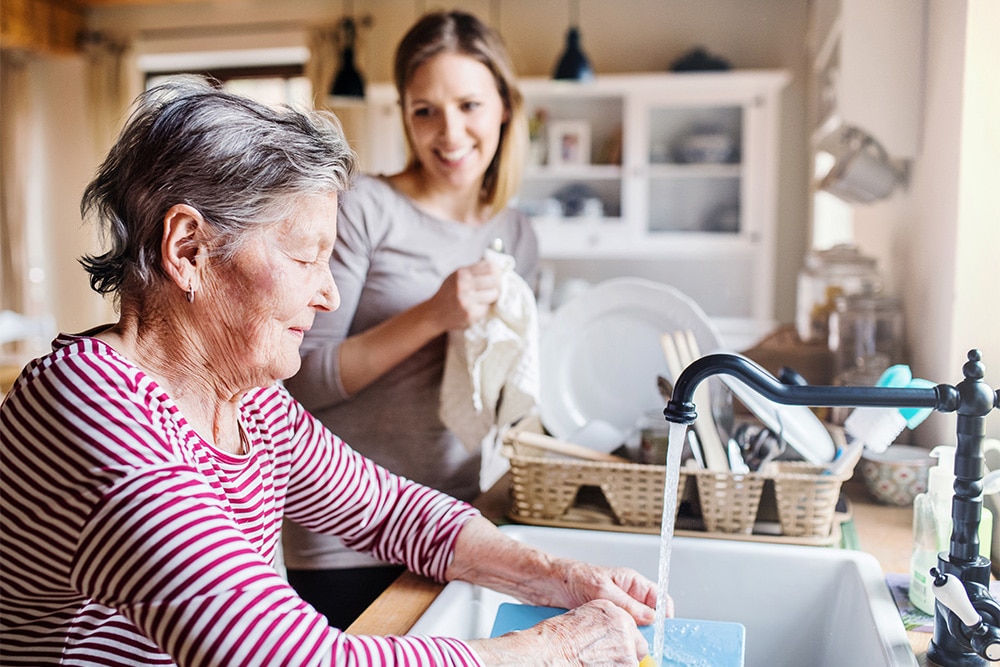 A caregiver helping an elderly woman with dishes in a bright kitchen
